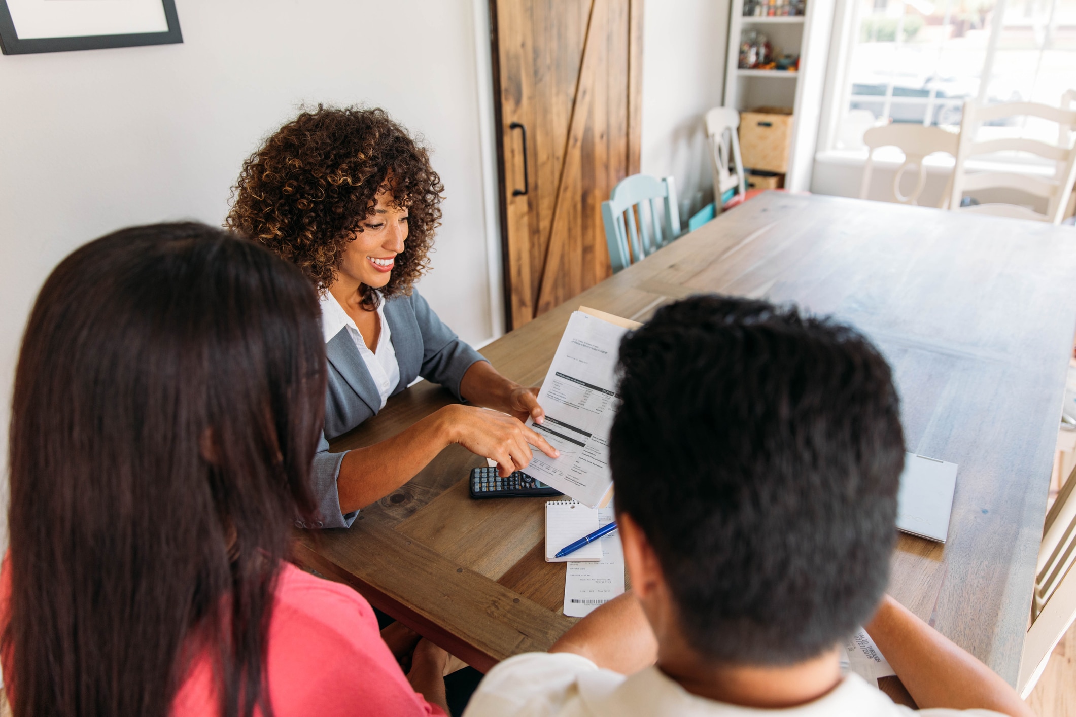 A female financial advisor sits with a millennial age married couple at a dining room table with laptop and financial reports helping with their monthly budget and investments. She is showing her clients how to read financial reports and monthly finances, pay taxes and save money for the future.